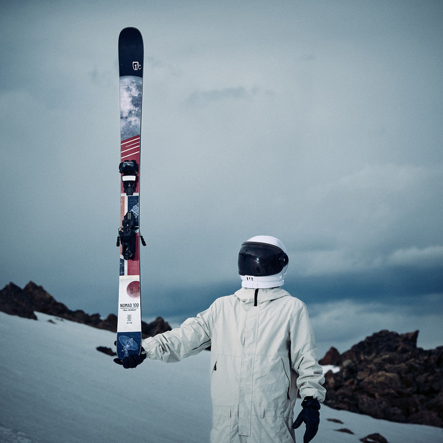 Wearing a white spacesuit and black gloves, an individual stands on a snowy slope holding an Icelantic Skis Nomad 100 upright with one hand; rocky terrain and a cloudy sky provide the backdrop.