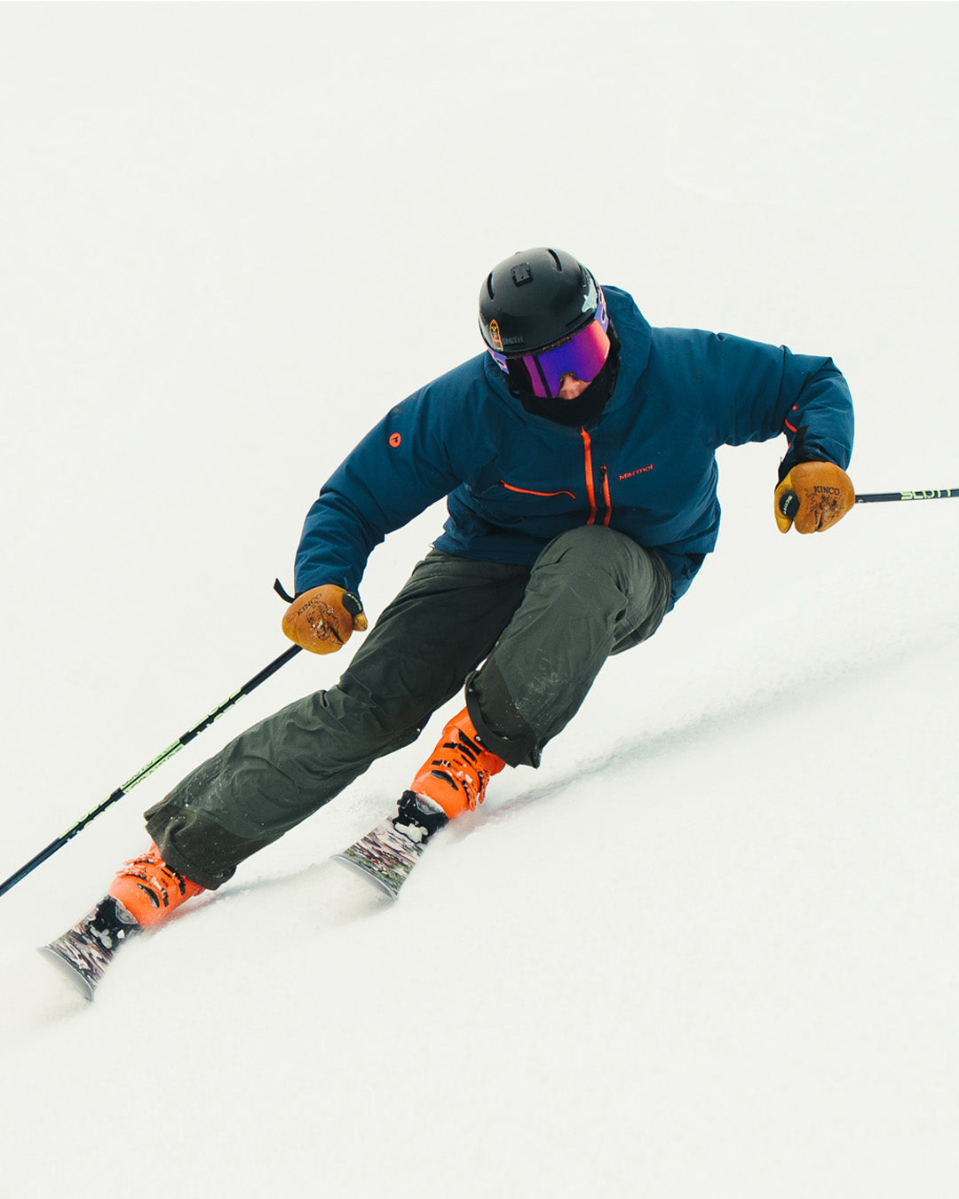 A skier in a blue jacket, orange gloves, and orange boots makes a sharp turn on fresh snow, wearing a black helmet and reflective goggles.