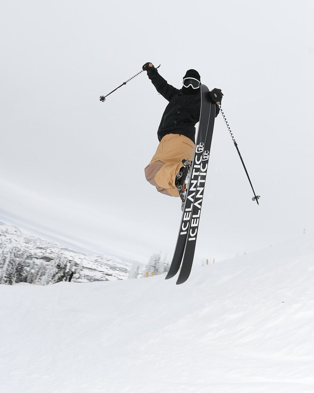 A skier wearing tan pants and a black jacket jumps in the air on a snowy slope, holding ski poles and skis labeled ICELANTIC, with snow-covered trees visible in the background.