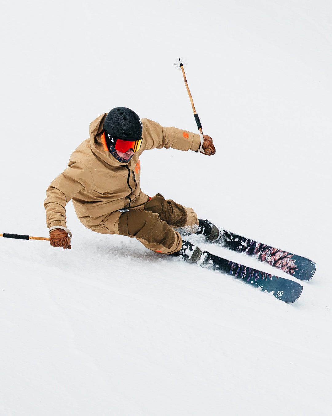 A skier in tan gear and a black helmet makes a sharp turn on snowy slopes, holding ski poles and wearing red goggles.