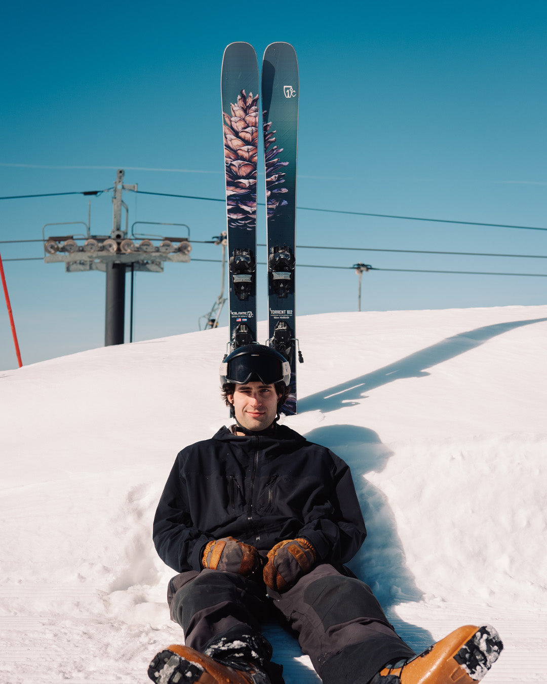 A skier in black gear sits on snow, wearing a helmet and goggles, with skis stuck upright behind him, blue sky and ski lift cables in the background.