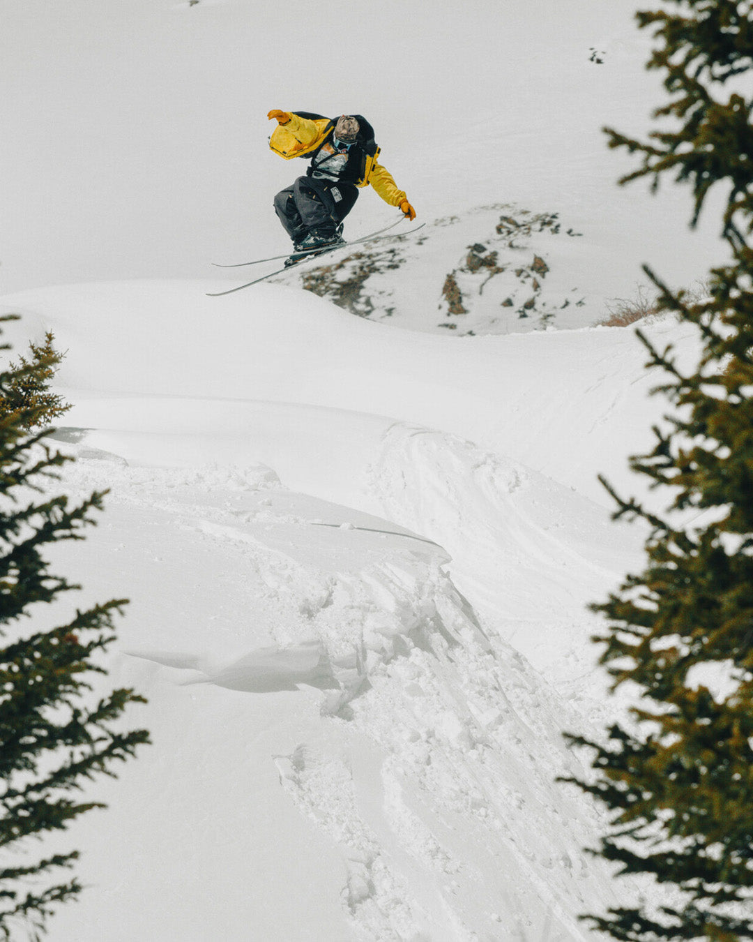 A skier in a yellow jacket jumps off a snowy ledge, surrounded by snow-covered slopes and trees on either side.