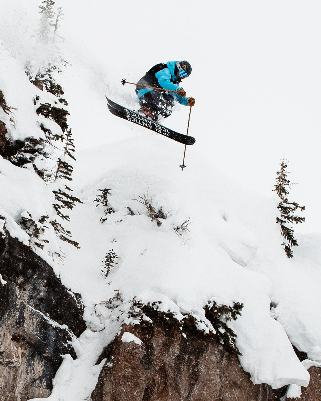 A skier in a blue jacket jumps off a snowy cliff, surrounded by snow-covered trees and rocky terrain.