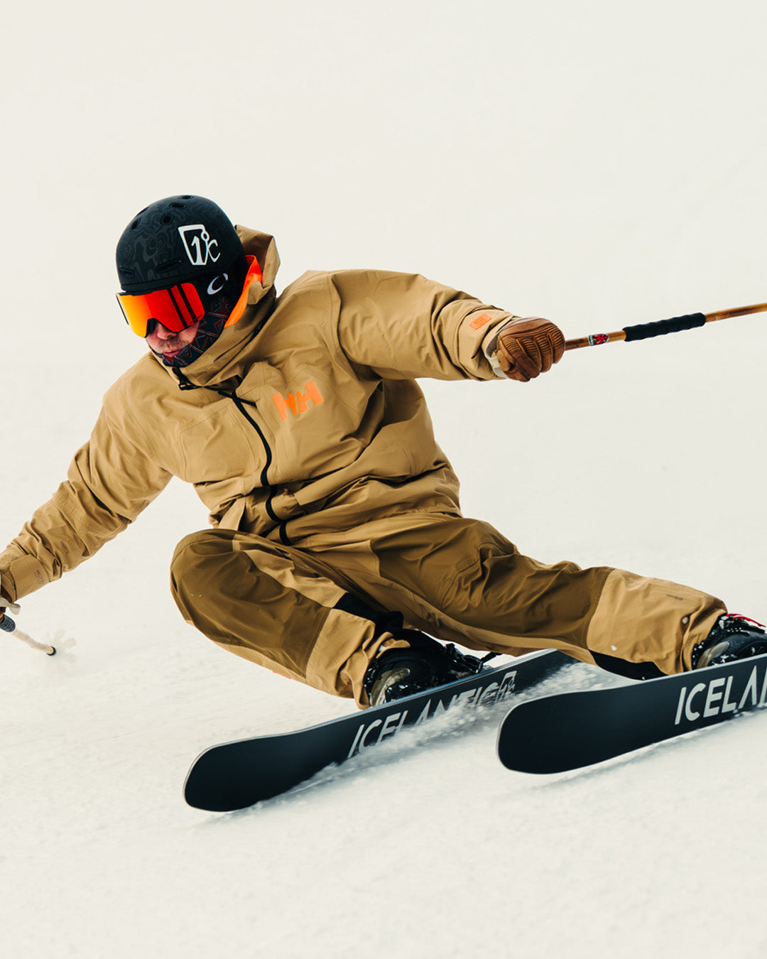 A skier wearing a tan jacket and pants, black helmet, and red goggles is turning sharply on snow, creating a spray as they carve with black skis labeled ICELANTIC..
