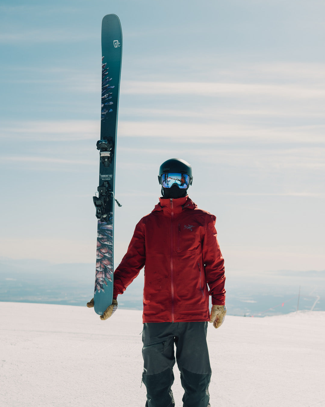 A skier in a red jacket, blue helmet, and ski goggles stands on snow, holding one ski upright with a snowy landscape and blue sky in the background.