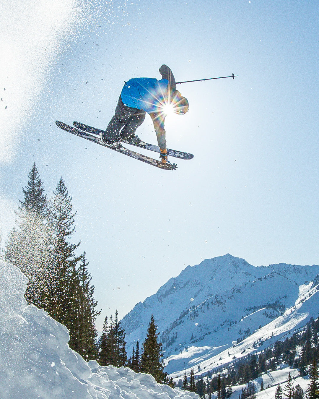 A skier in mid-air performs a trick above snowy slopes with mountains and trees in the background. Sunlight shines through the skiers arm.
