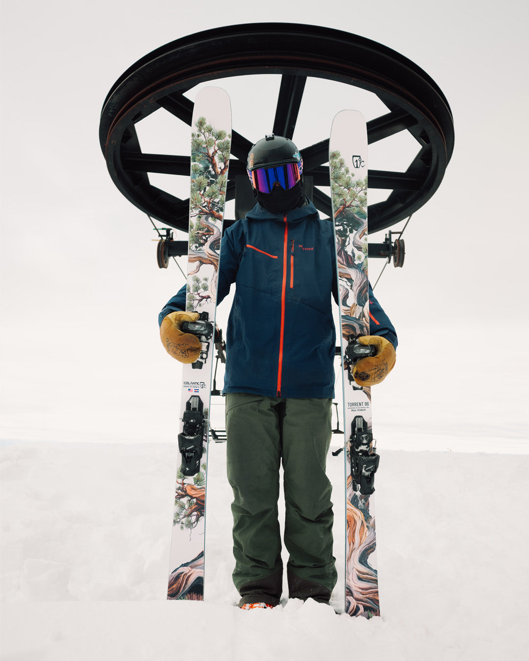 A skier wearing goggles and a helmet stands in the snow holding patterned skis upright, with a ski lift wheel above their head against a cloudy sky.