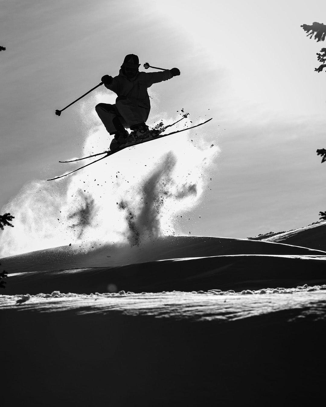 A skier is captured mid-air during a jump, silhouetted against the sky with snow spraying beneath them. The scene is black and white, highlighting the skiers dynamic movement and the snowy landscape.