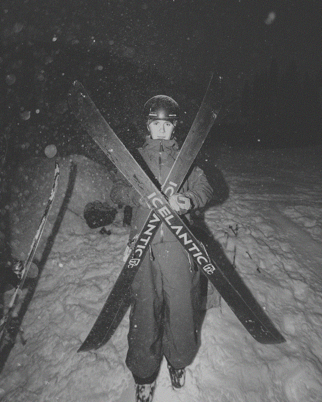 A person in winter gear stands in the snow at night, holding two crossed skis labeled ICELANTIC. Snow falls around them, and the scene is lit by a bright light.