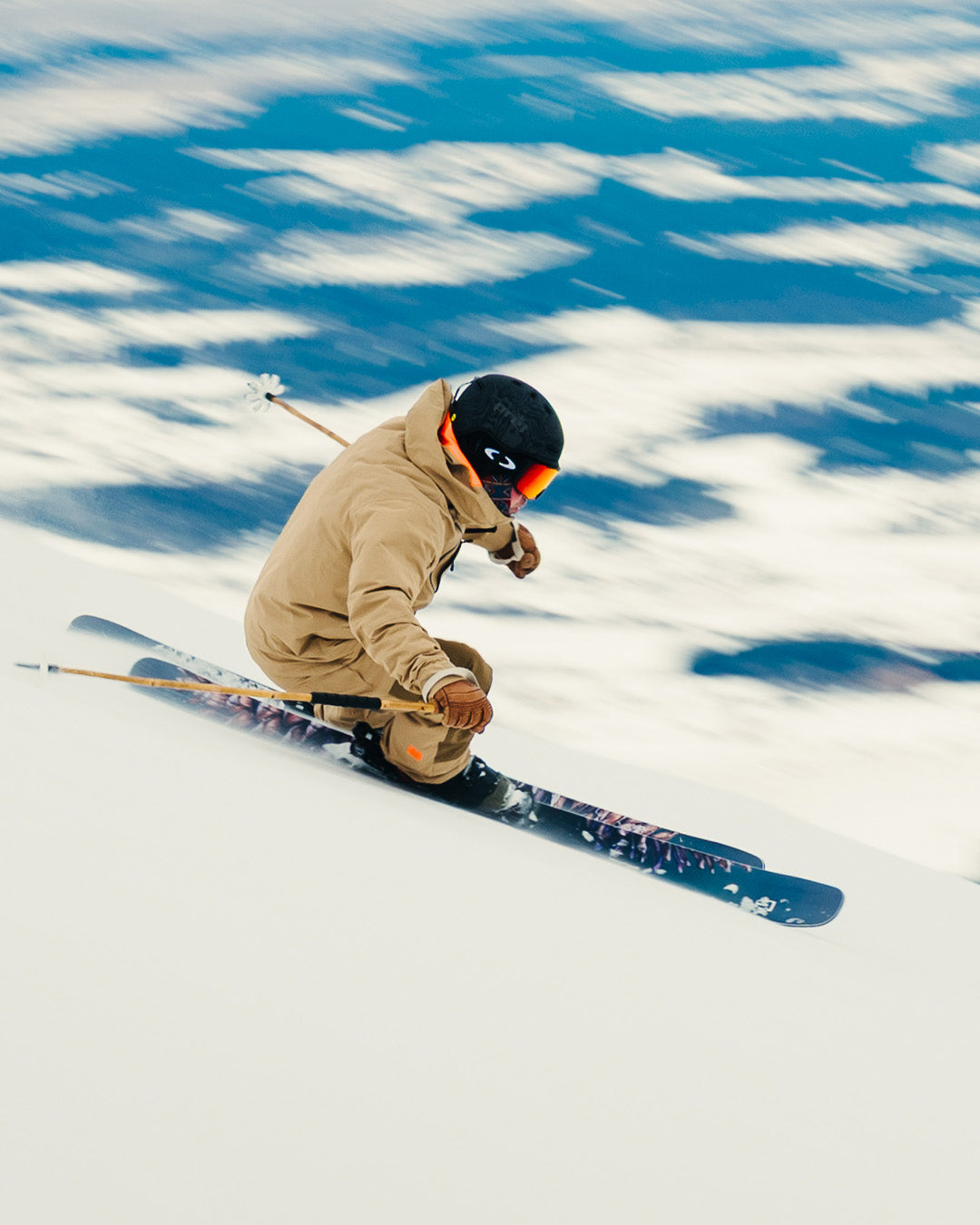 A skier in tan gear and a black helmet speeds downhill on a snowy slope, with blurred blue and white scenery in the background suggesting fast movement.