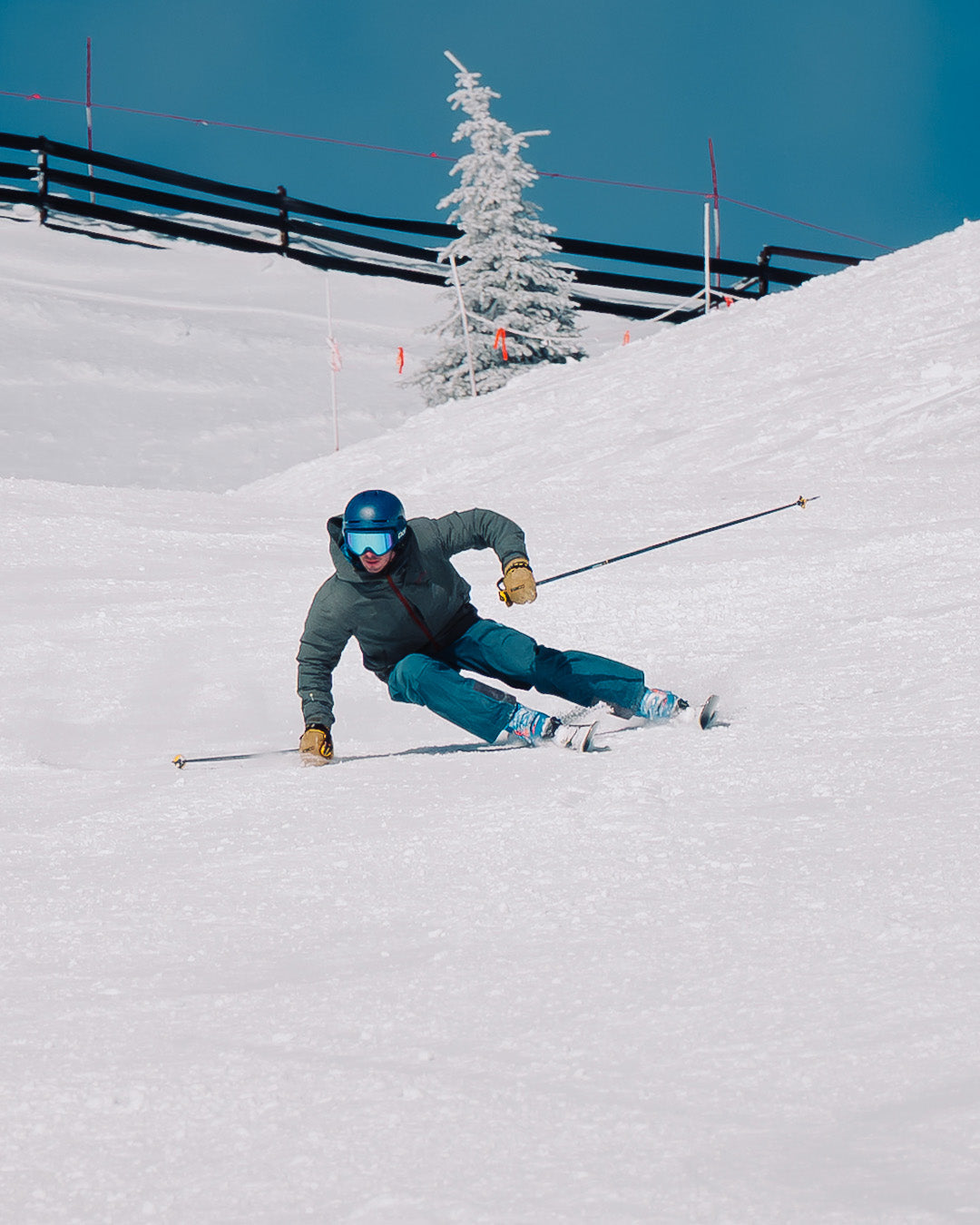 A skier in a blue helmet and green jacket makes a sharp turn on a snowy slope, holding ski poles. A frosted tree and fence are visible in the background under a clear blue sky.