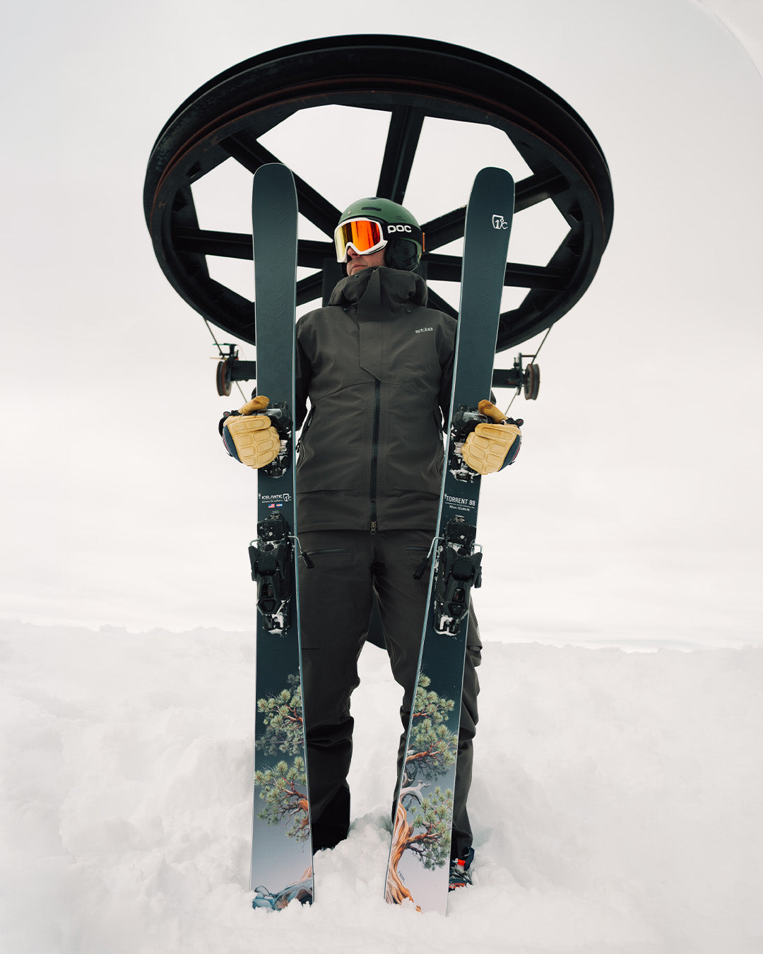 A skier in black gear holds two skis upright, standing in snow beneath a ski lift wheel. The skis have a tree design near the base. The skier wears goggles, gloves, and a helmet.