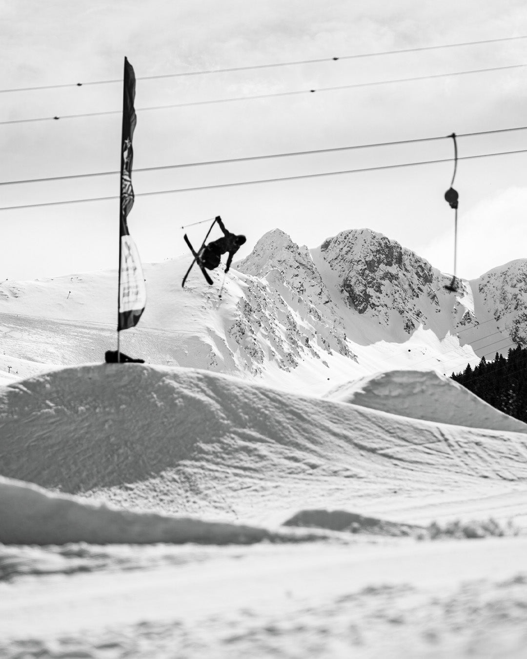 A skier performs an aerial trick above a snowy slope with mountains in the background. The image is black and white, and ski lift cables are visible overhead.