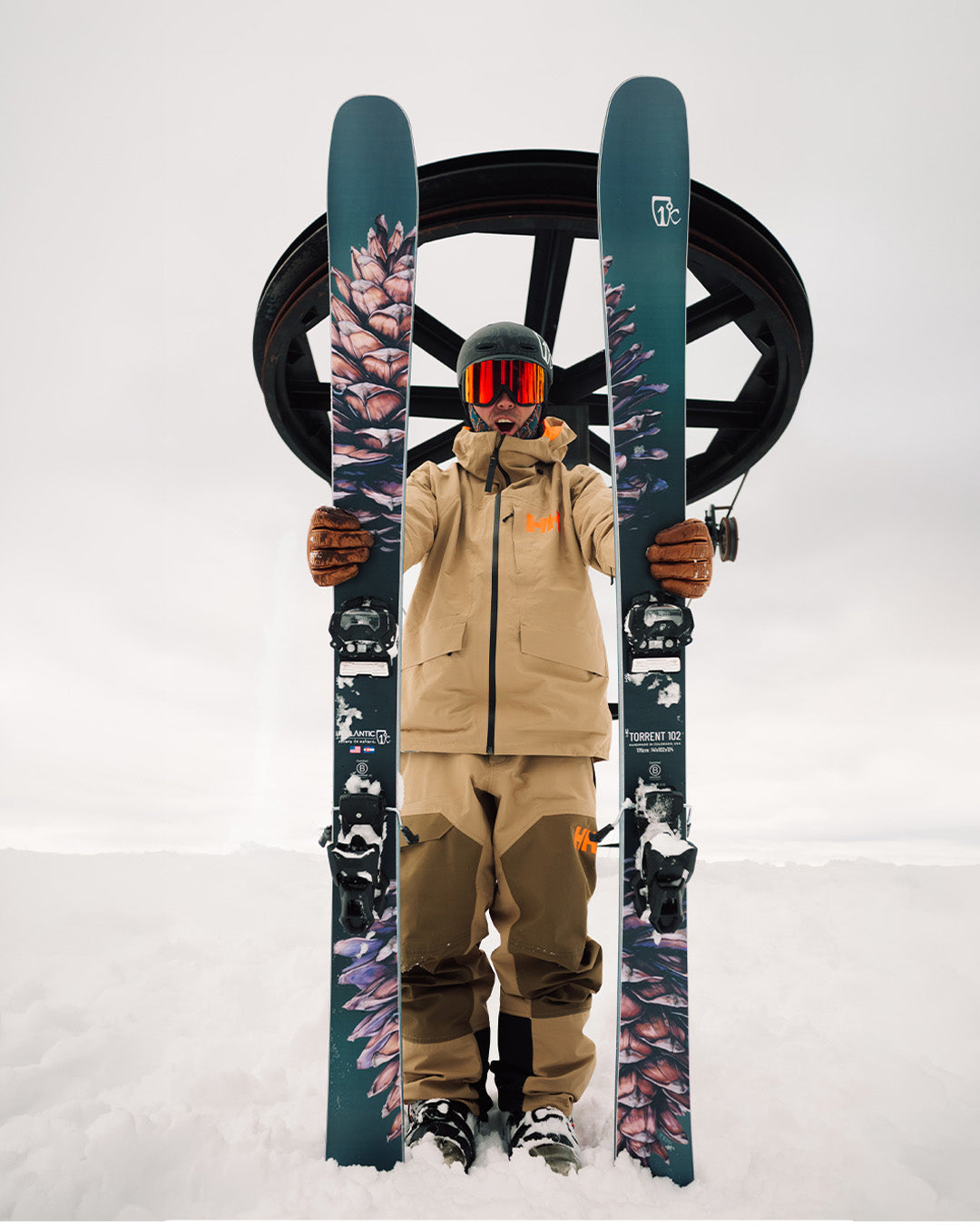 A person in tan ski gear stands in snow, holding two blue skis with feather designs vertically in front of a ski lift wheel. The persons face is covered by goggles and a helmet.