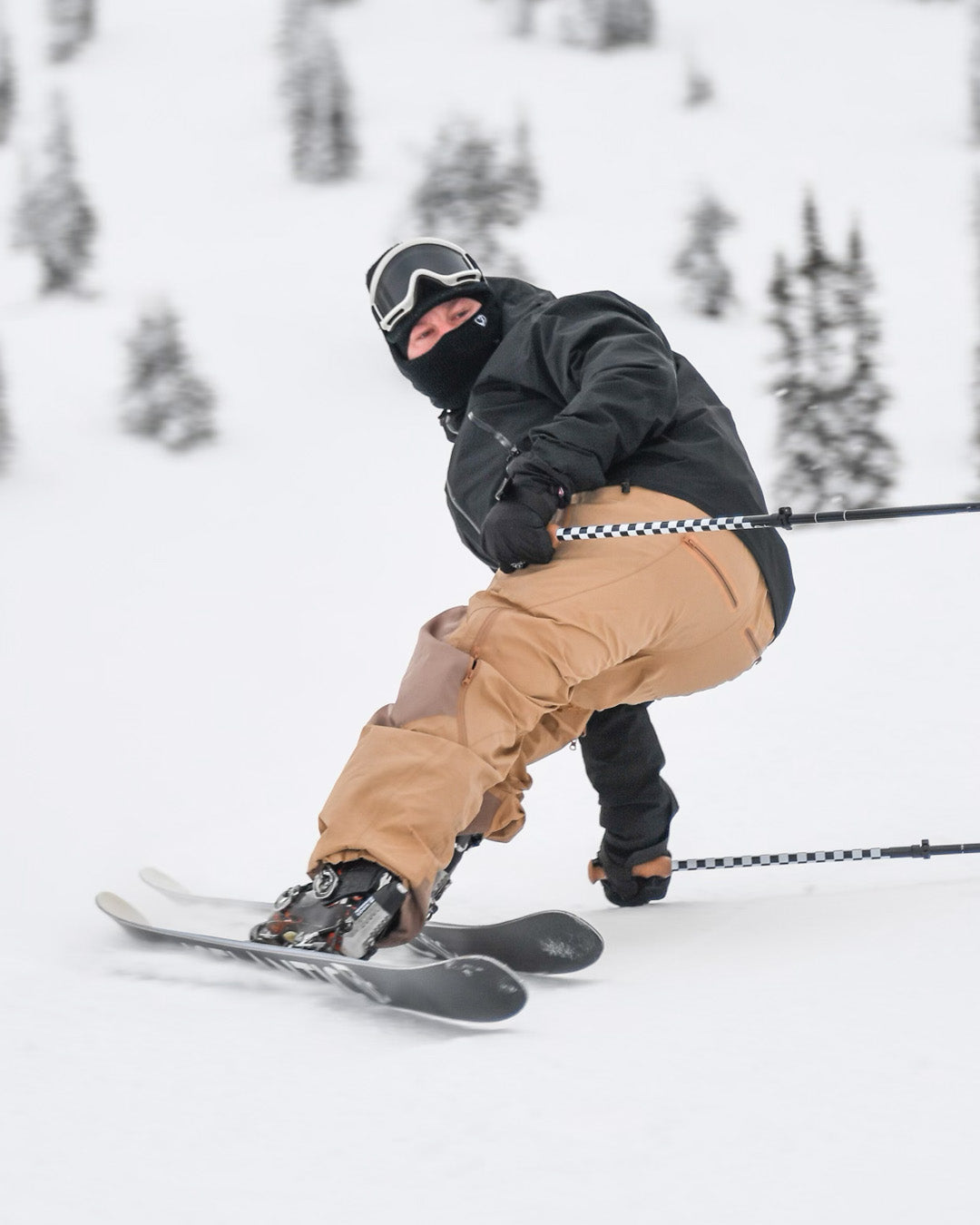 A skier in tan pants and a black jacket, mask, and goggles makes a sharp turn on snowy slopes with pine trees in the background.