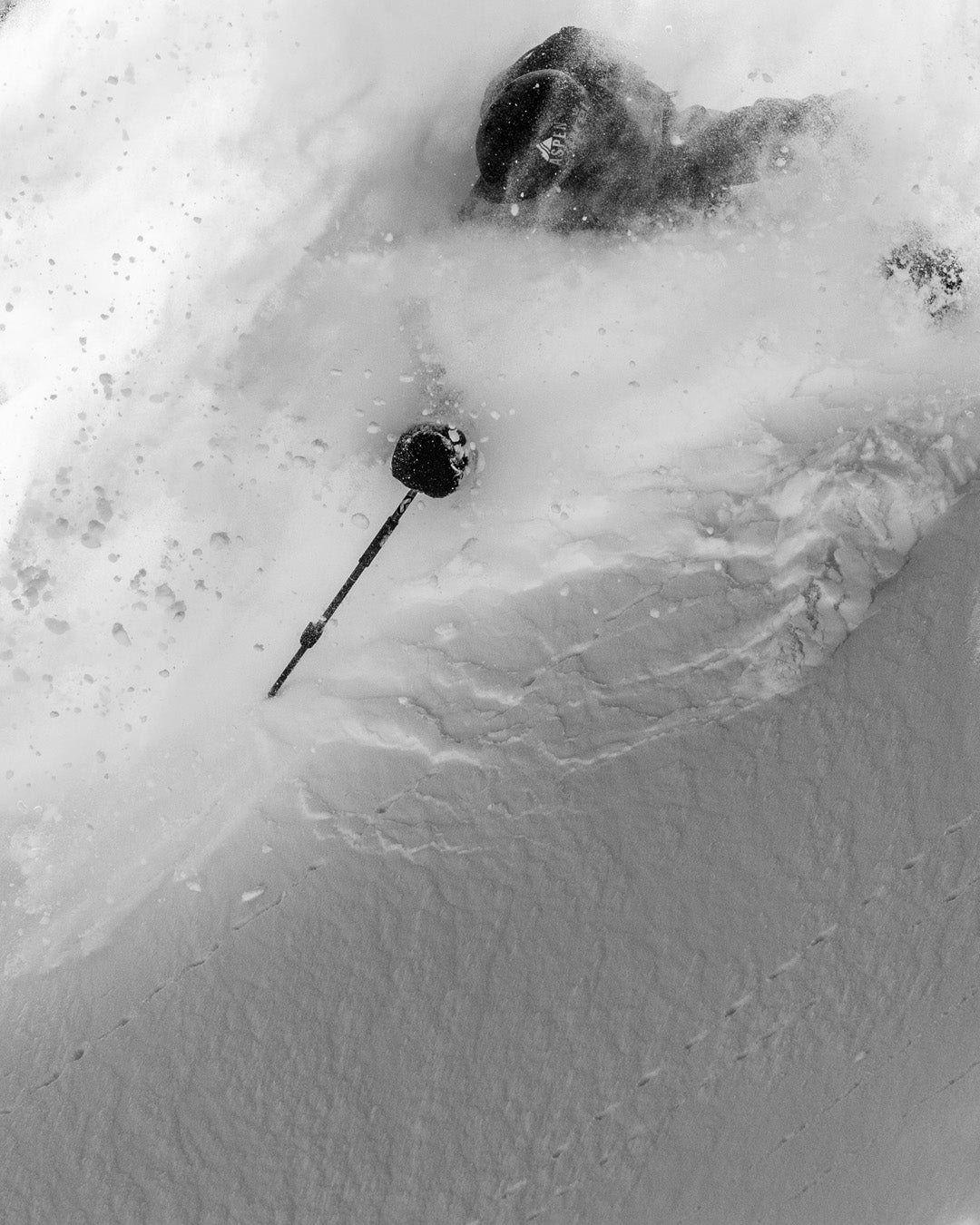 A skier, mostly covered in snow, moves downhill with only part of their body and a ski pole visible amid the deep powder. The image is in black and white.