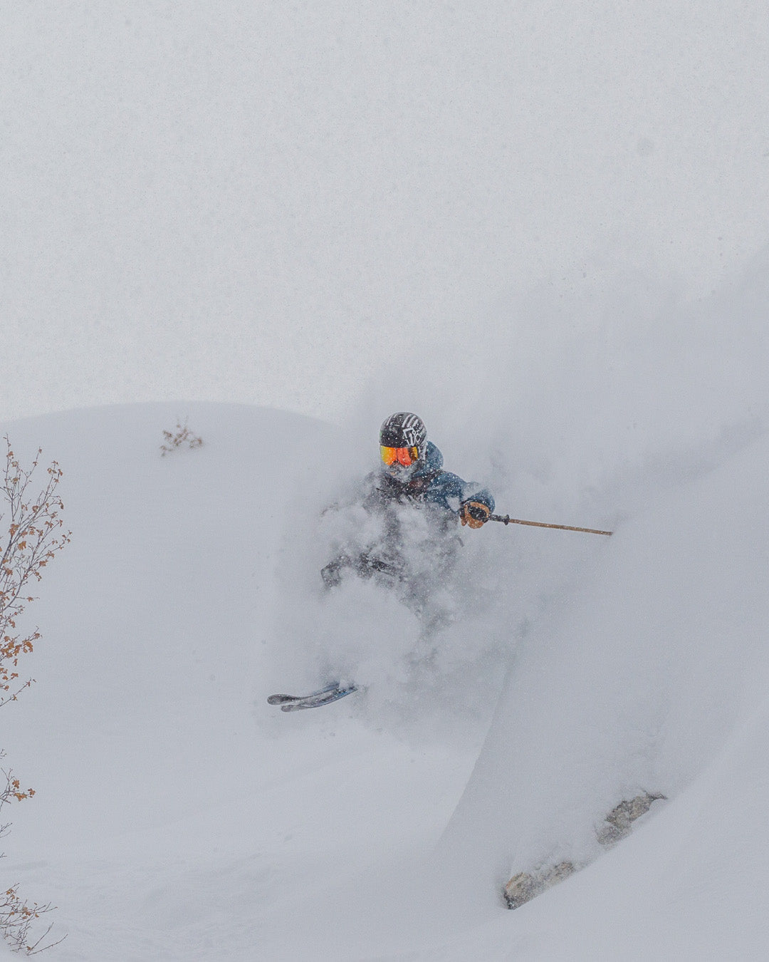 A skier wearing a helmet and goggles speeds downhill through deep powder snow, partially obscured by a burst of snow in a wintry landscape.