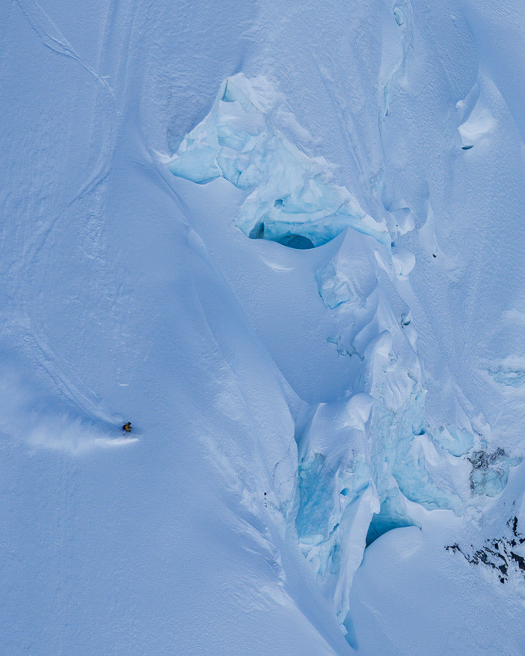 A skier descends a steep, snow-covered mountain slope near ice formations and crevasses, leaving a trail in the fresh snow.