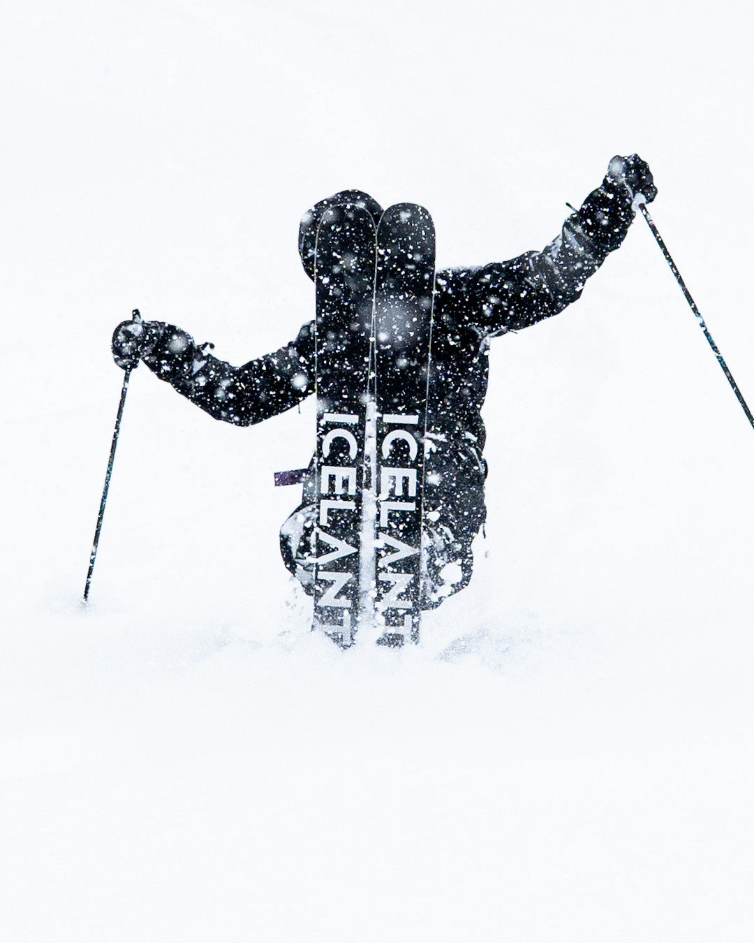 A skier in heavy snow, holding ski poles and wearing skis labeled ICELANT, is seen from behind, surrounded by white snow and snowfall, blending into the wintry landscape.