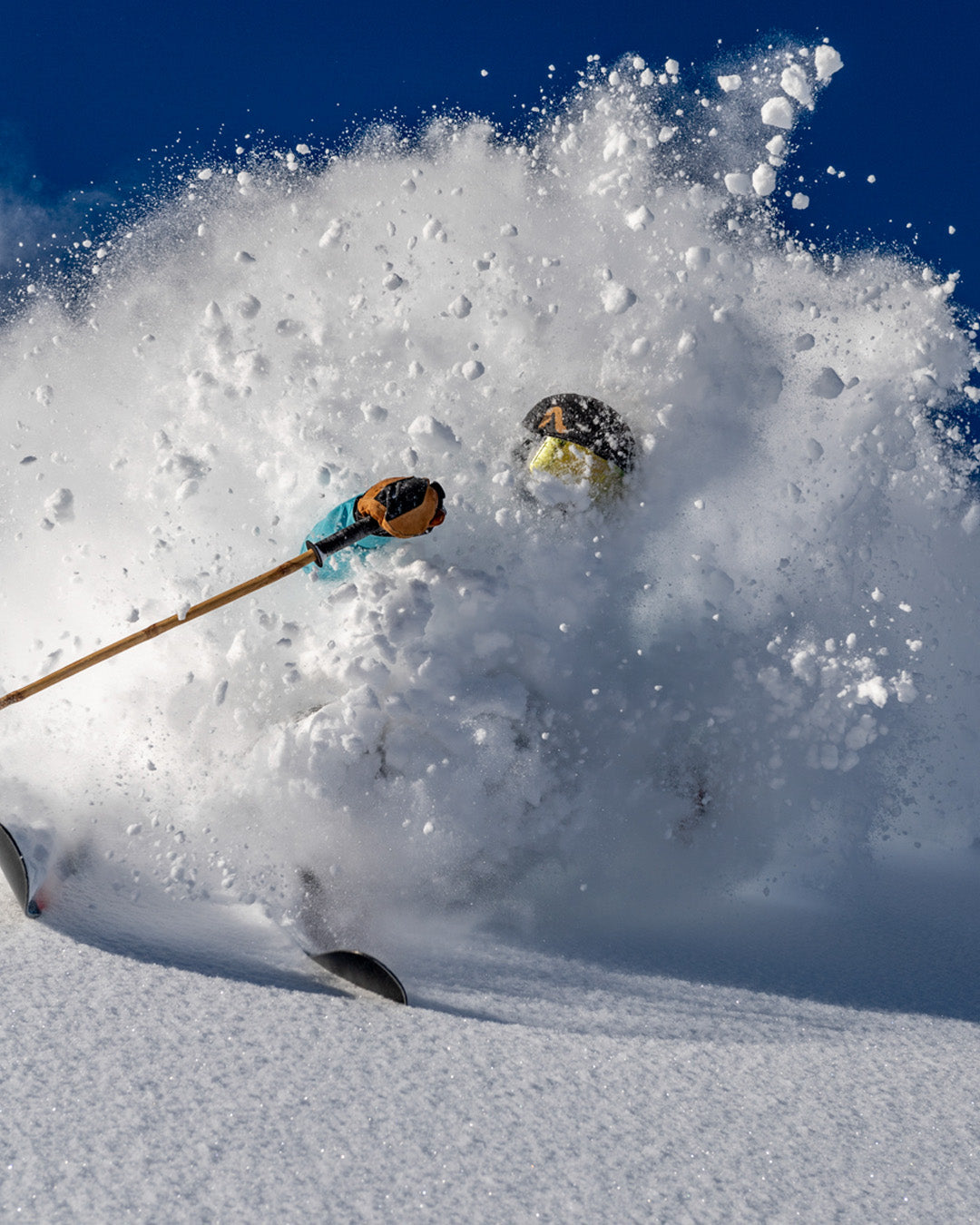 A skier bursts through fresh powder snow, creating a massive spray that nearly obscures them, with only their goggles, pole, and ski tips visible against a clear blue sky.