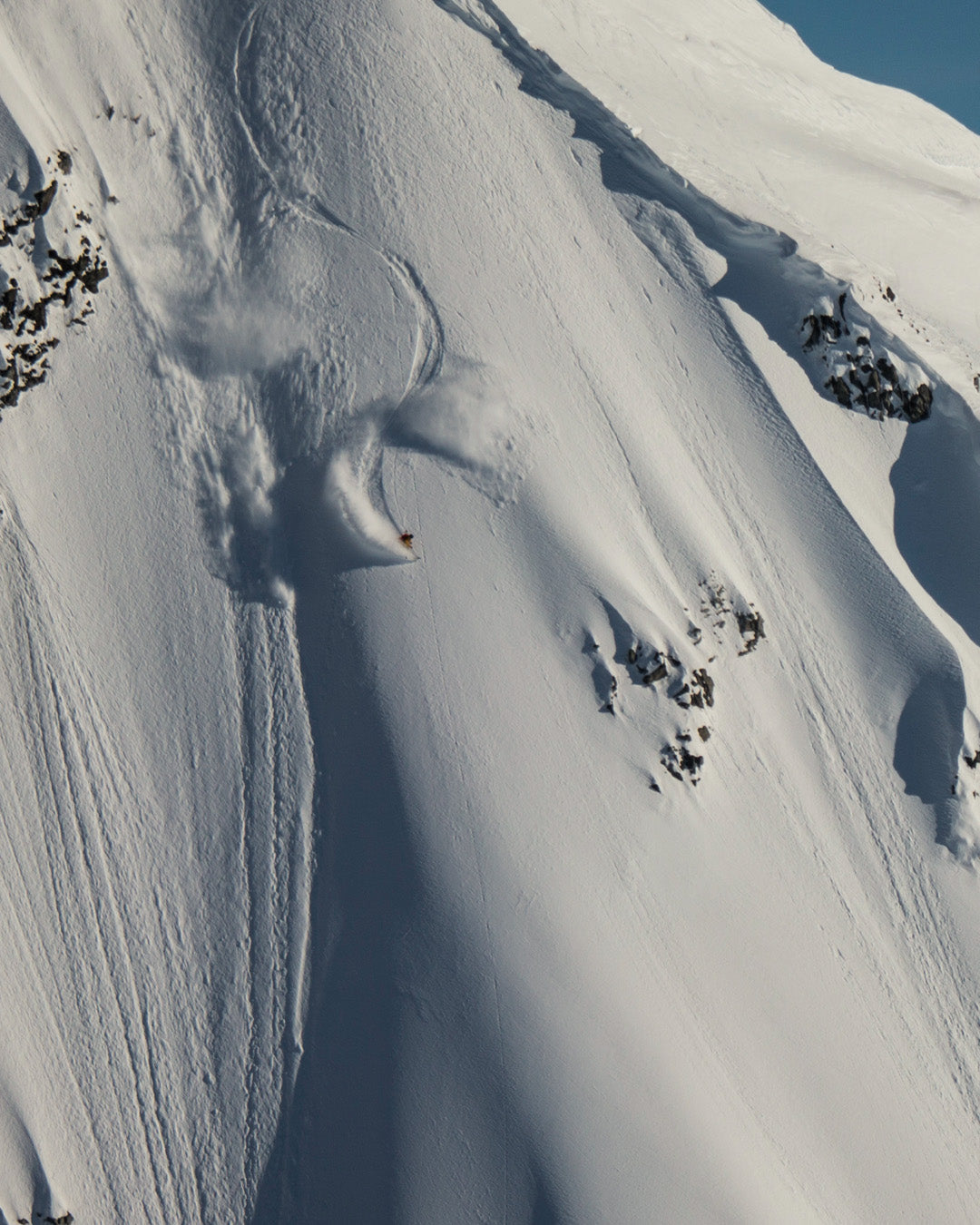 A skier makes fresh tracks down a steep, snow-covered mountain slope, carving through powder near rocky outcrops under a clear sky.