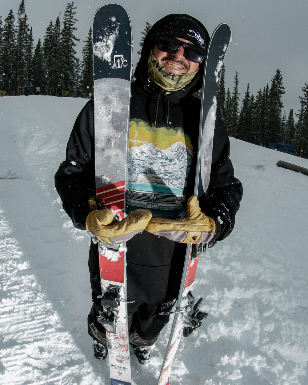 A smiling skier in sunglasses and gloves holds two skis upright on a snowy slope, surrounded by pine trees and winter scenery.