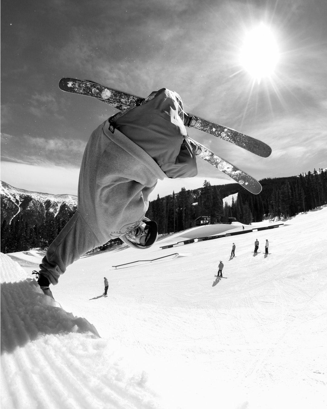 A skier mid-air performs a trick on a snowy slope under a bright sun, with trees, mountains, and several people in the background. The image is in black and white.