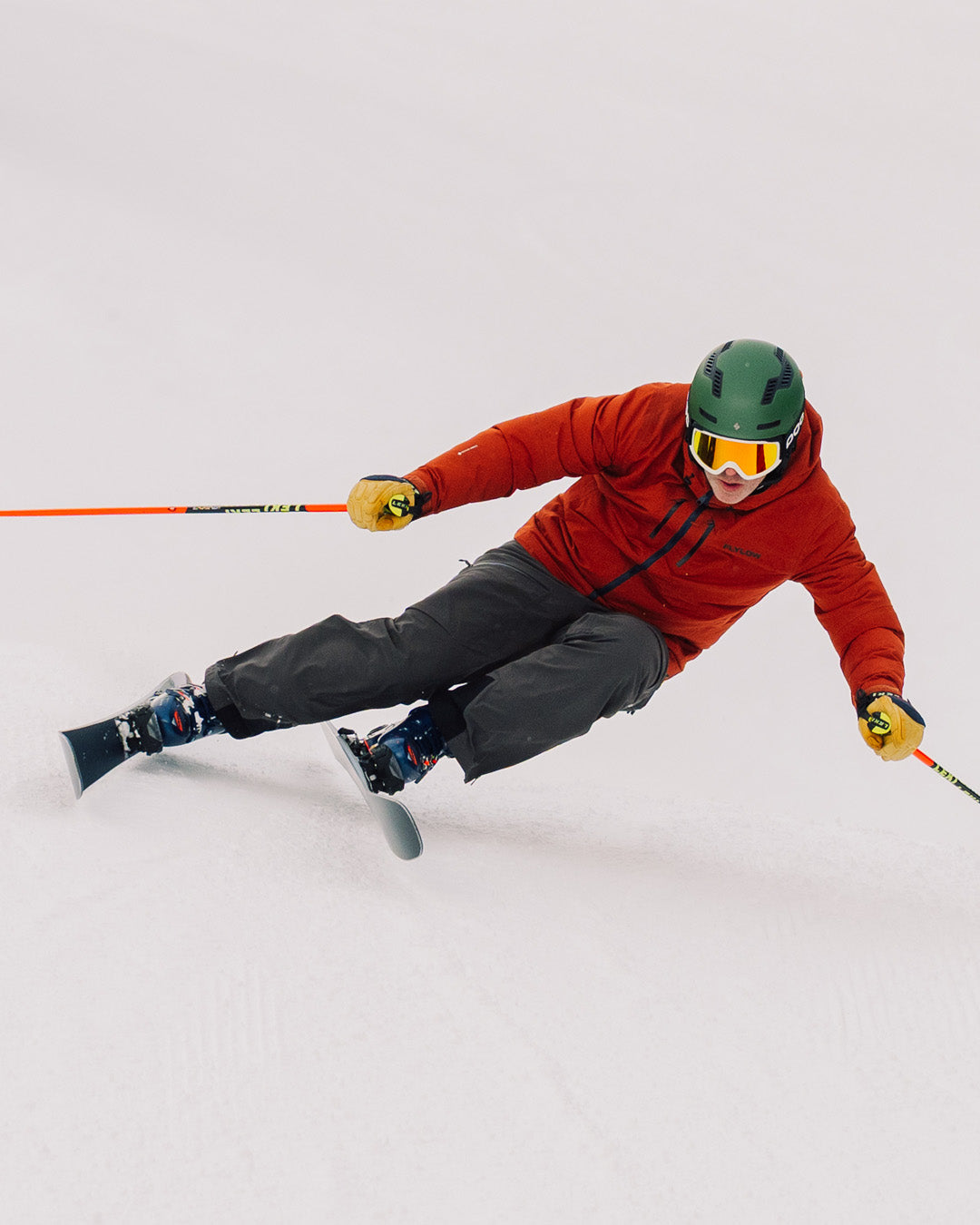 A skier in a red jacket and green helmet makes a sharp turn on a snowy slope, holding ski poles and wearing yellow goggles.