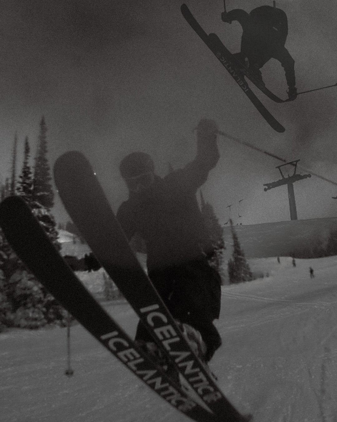 Two skiers perform jumps on a snowy slope; one is airborne in the background, while the other is close to the camera. Pine trees and a ski lift appear in the distance. The scene is in black and white.