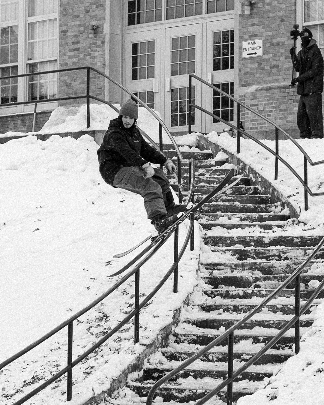 A skier slides down a handrail beside snowy steps outside a building, while a person films the action nearby.