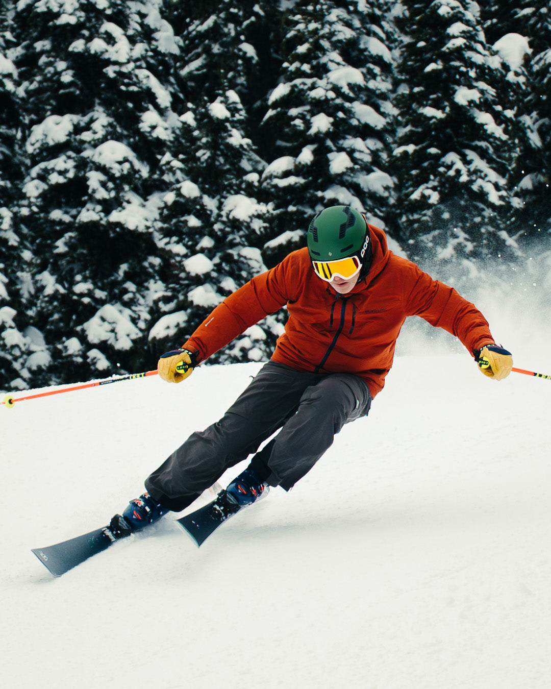 A skier in a green helmet and orange jacket carves down a snowy slope, surrounded by snow-covered trees, holding ski poles in both hands.