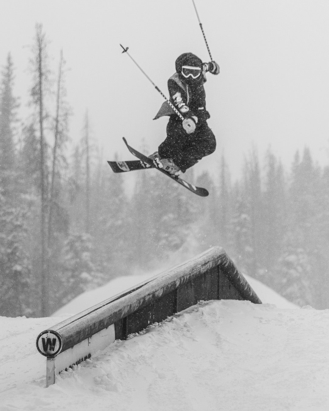 A skier in midair performs a trick over a rail in a snowy, forested area. The skier is wearing goggles, a helmet, and winter gear. The image is in black and white.