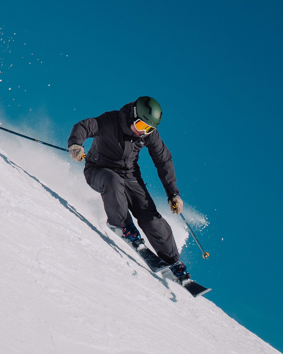 A skier in a dark outfit and green helmet speeds down a snowy slope, leaning forward with ski poles in hand against a clear blue sky.