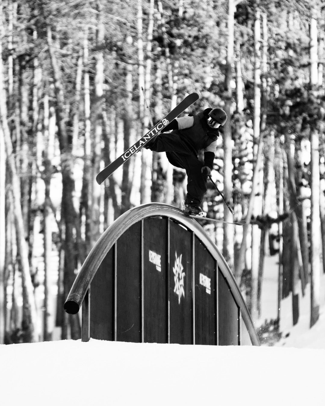 A skier performs a trick on a curved rail in a snowy terrain park, holding one ski with one hand, surrounded by tall, leafless trees.