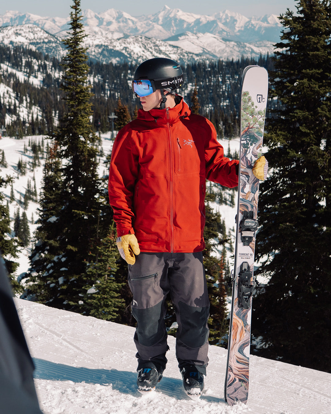 A skier in a red jacket, black helmet, and goggles stands holding skis on a snowy mountain slope with pine trees and distant peaks in the background.