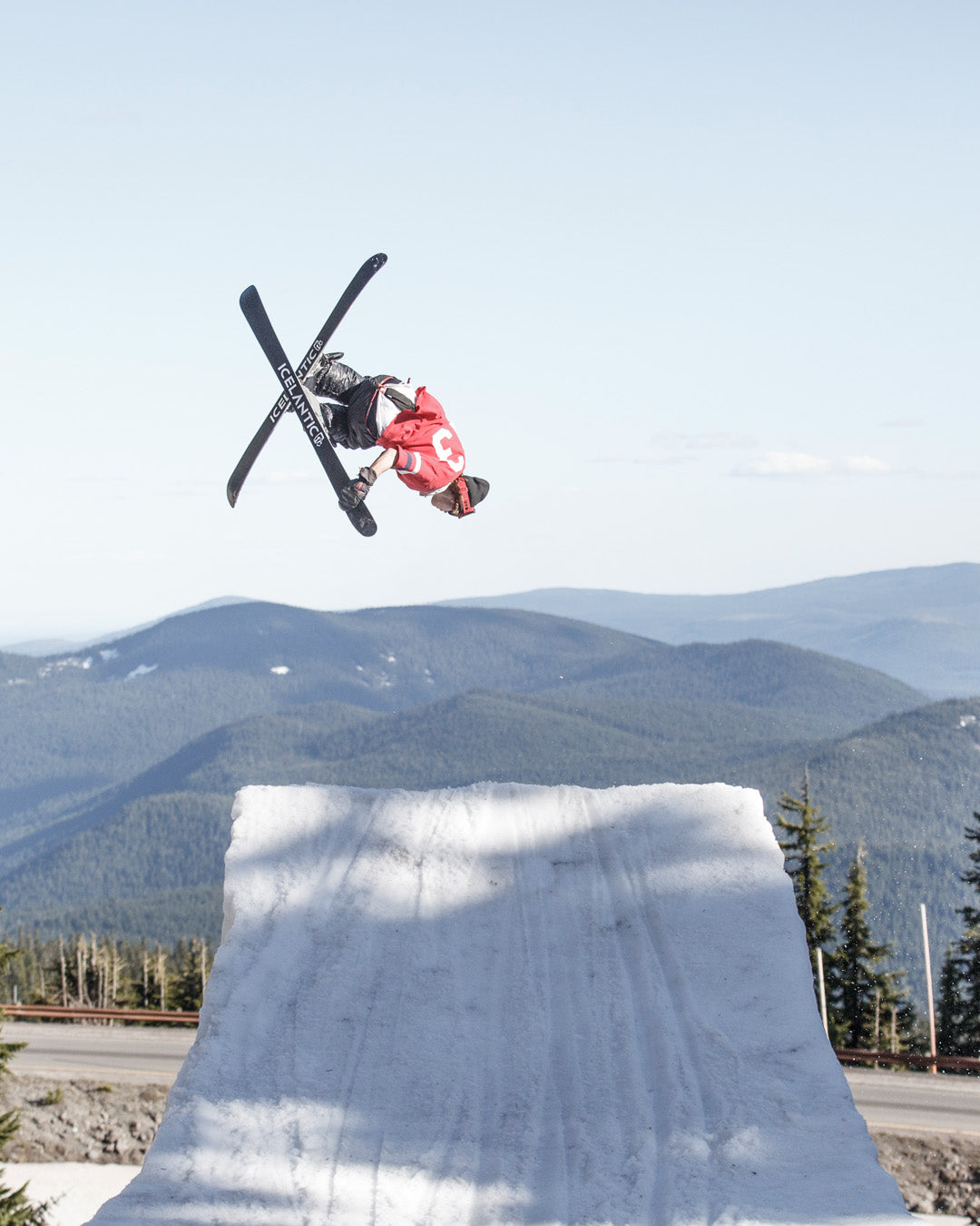 A skier in midair performs a trick above a snowy ramp, with mountains and trees in the background under a clear sky.