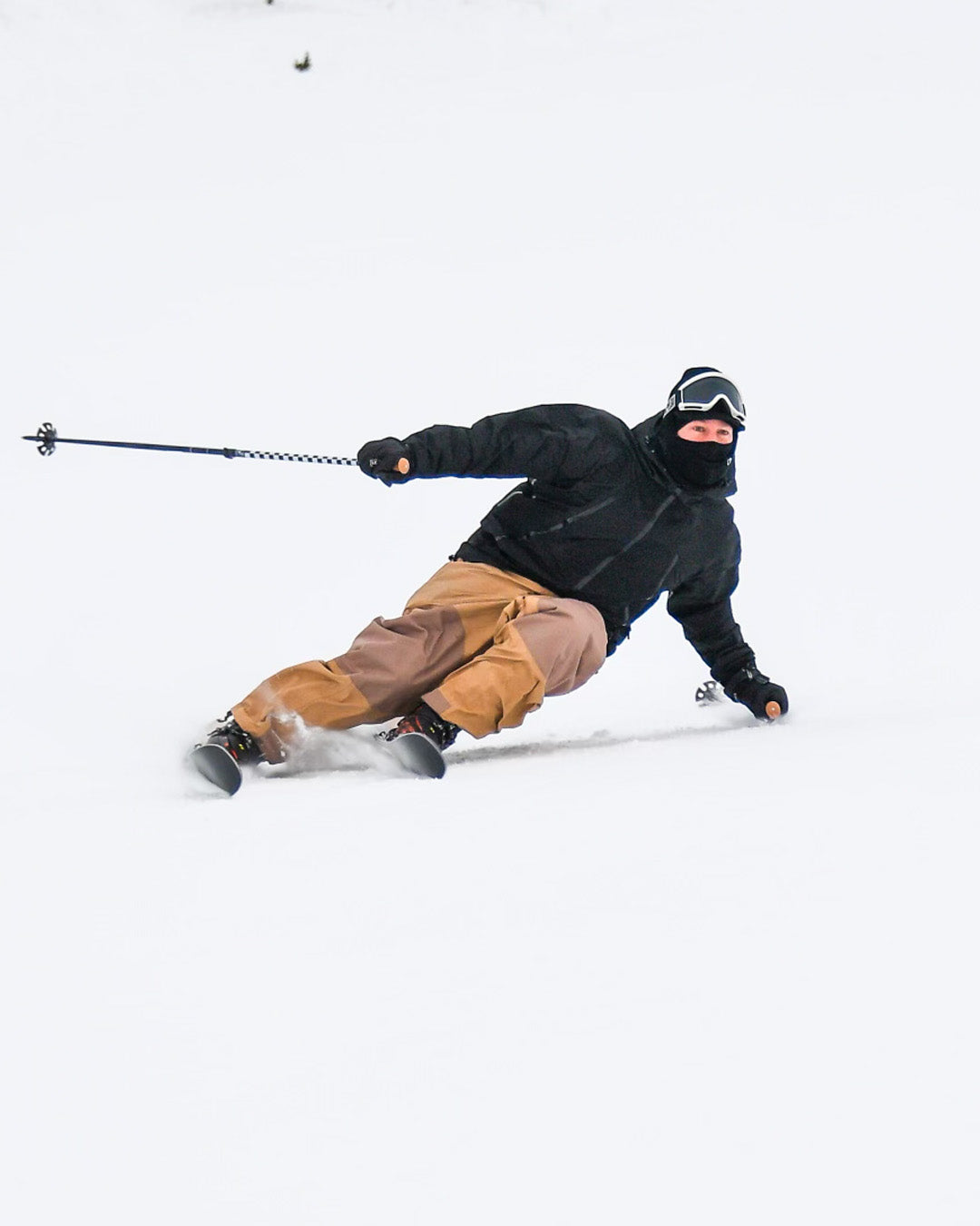 A skier in brown pants and a black jacket carves sharply on snow, leaning close to the ground with ski poles angled behind, wearing goggles and a face covering.