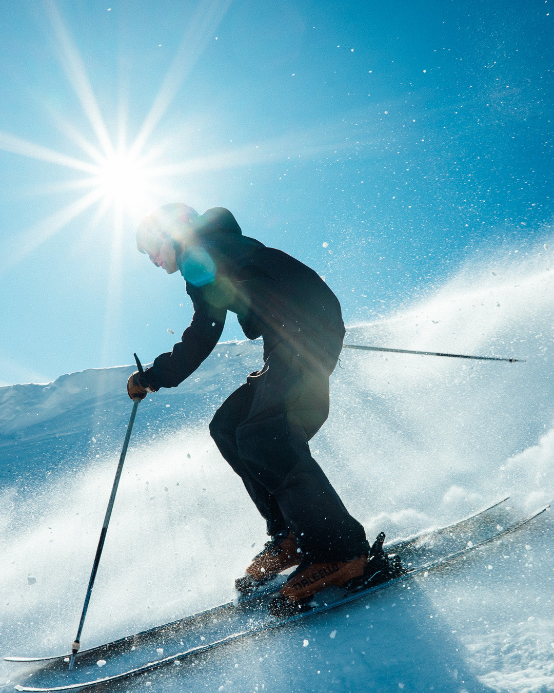 A skier dressed in dark clothing skis downhill on a sunny, snowy slope with bright sunlight and a spray of snow in the background.