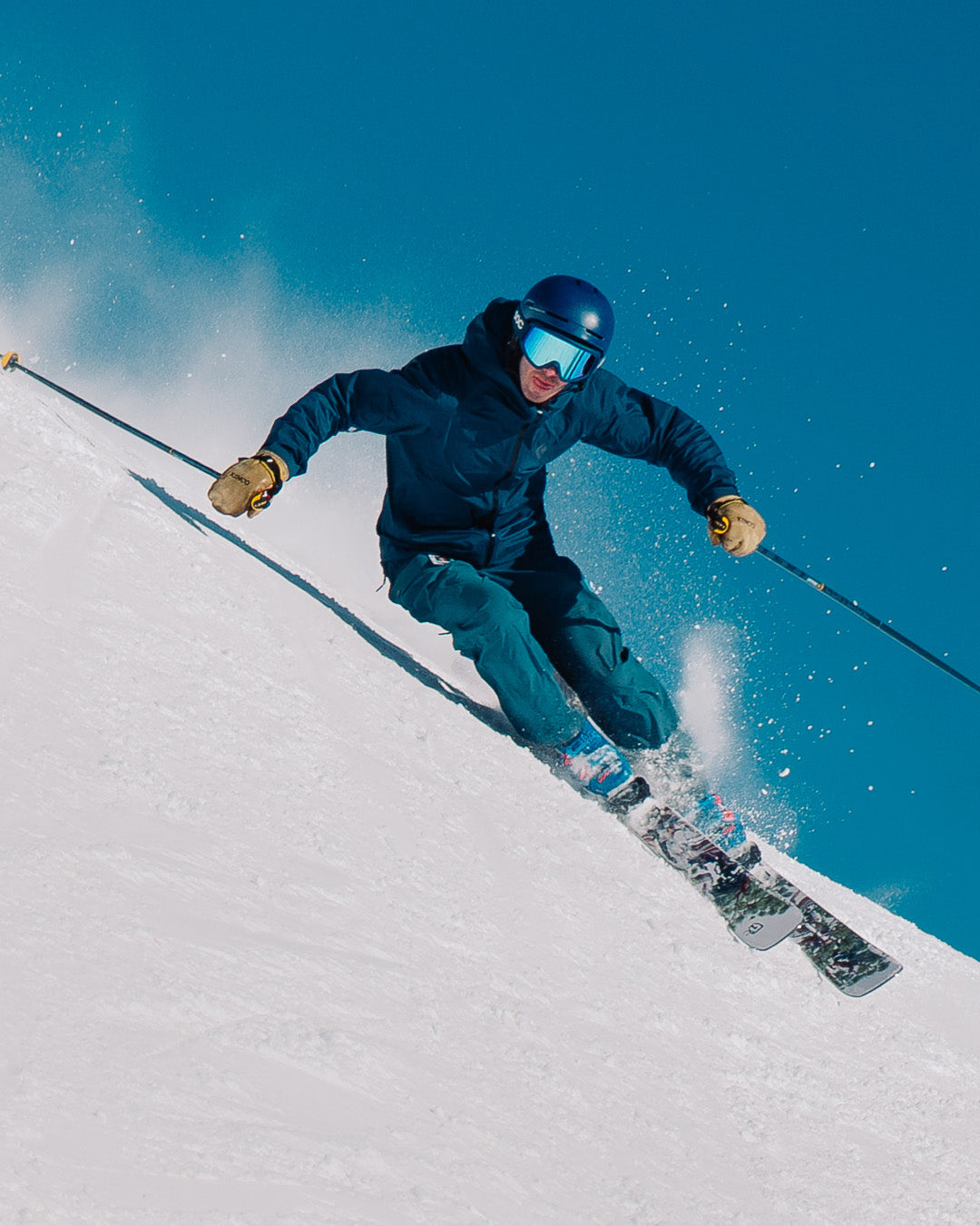 A skier in blue gear and helmet descends a snowy slope at high speed, kicking up snow against a clear blue sky.
