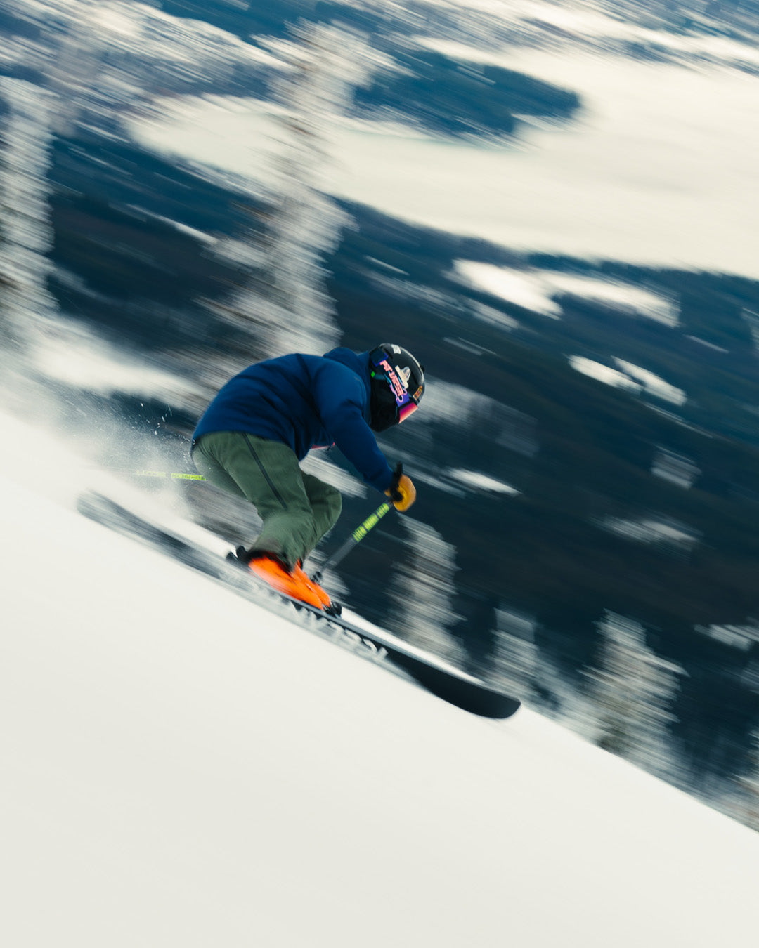 A skier in a blue jacket and green pants speeds downhill on a snowy slope, creating a blur effect in the background.