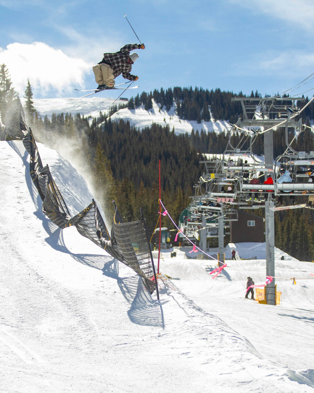 A skier in mid-air performs a trick above a snow ramp near a ski lift, with mountains and trees in the background under a clear blue sky.