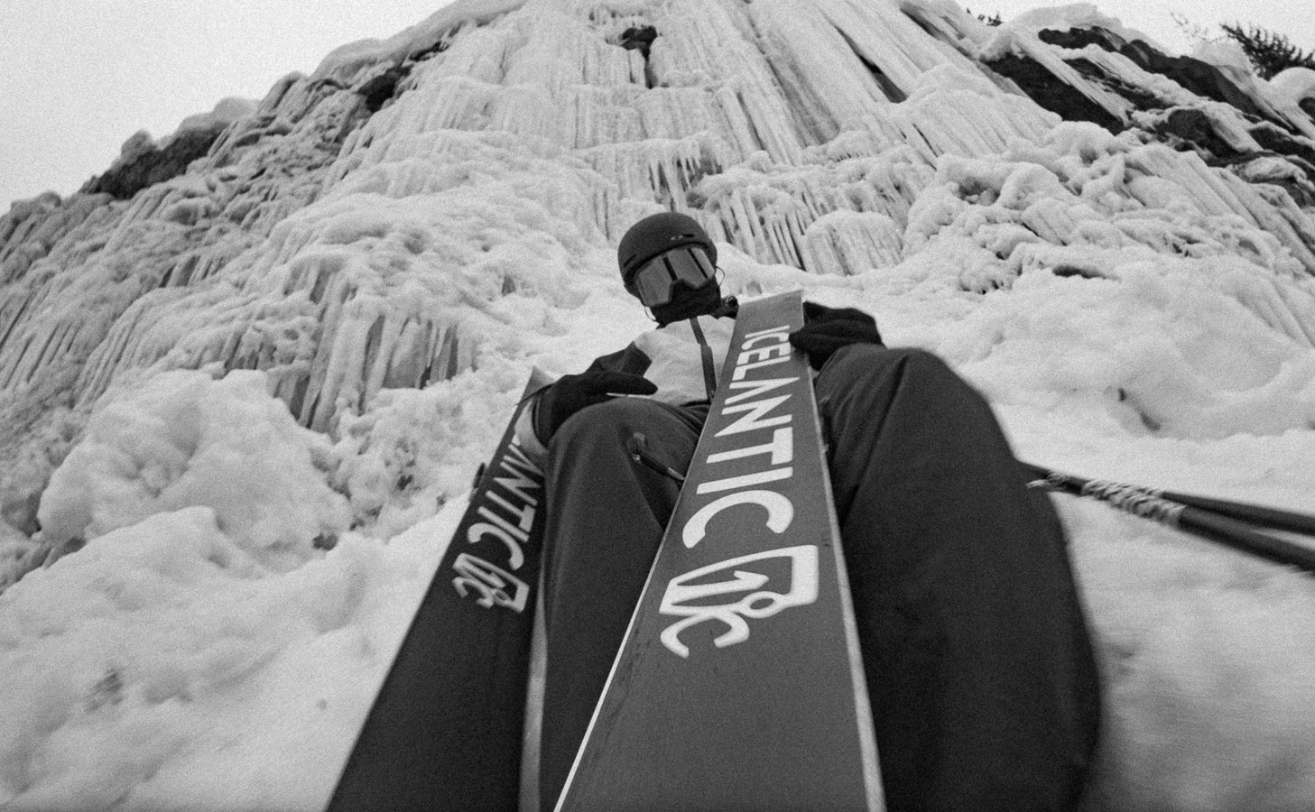 A skier in a helmet and goggles sits at the base of a snowy, icy slope, holding ski poles. The photo is black and white, with the Icelantic Skis Nomad 106 brand visible on both skis.