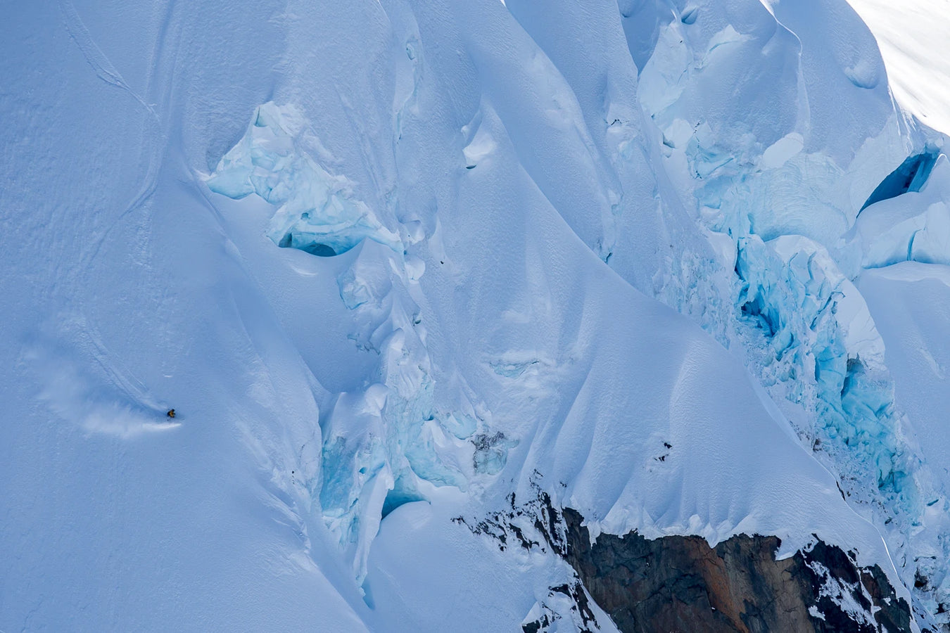 A skier on the Icelantic Skis Nomad 112 carves down a steep, snow-covered mountain slope dotted with blue crevasses and rocky outcrops.