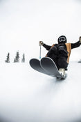 Clad in winter gear and goggles, a person rides the Icelantic Skis Nomad 100 Whiteout swiftly down a snowy slope, snow spraying around them as evergreens stand tall in the background.