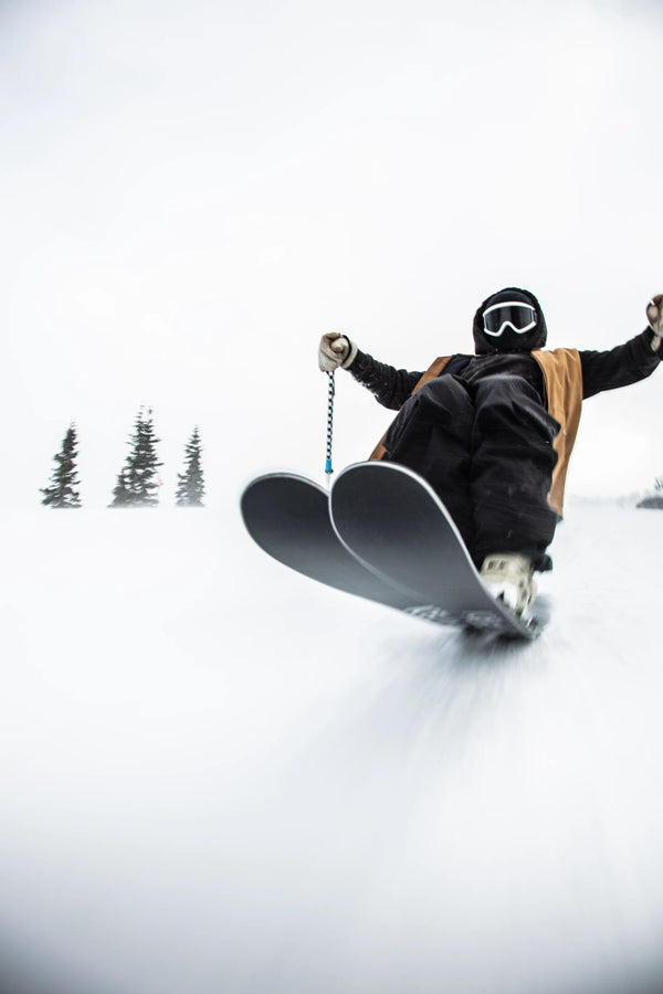 Clad in winter gear and goggles, a person rides the Icelantic Skis Nomad 100 Whiteout swiftly down a snowy slope, snow spraying around them as evergreens stand tall in the background.