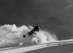 Clad in dark gear, a skier rides the Icelantic Skis Nomad 112 Whiteout downhill, carving through deep powder and sending up a billowing spray. Captured in black and white, the dynamic scene partially veils the skier behind a cloud of snow.