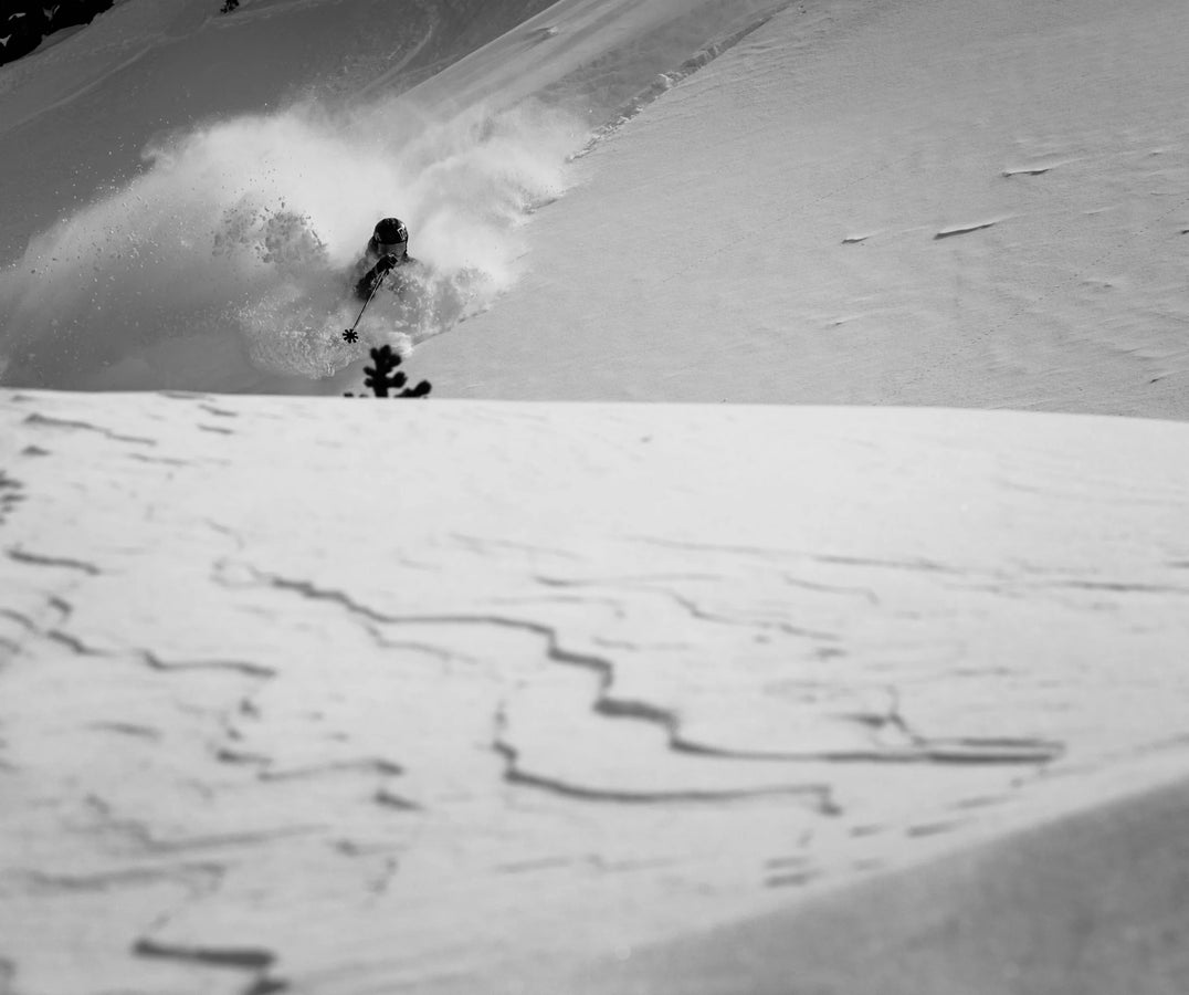 A skier on Icelantic Skis’ Nomad 112 Whiteout carves through deep powder snow on a mountain slope, creating a spray behind them. The black and white image highlights the snow patterns in the foreground.