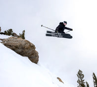 A skier outfitted in dark gear and goggles launches off a snowy slope on Icelantic Skis’ Maiden 102, ski poles extended wide, with trees and rocks scattered below beneath a cloudy sky.
