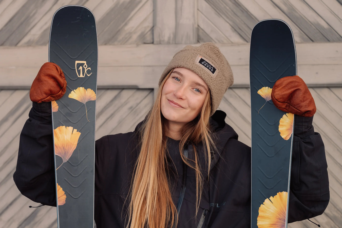 A woman in a tan beanie, brown gloves, and a dark jacket smiles while holding Icelantic Skis Tempest 88 skis with yellow flower designs, standing in front of a wooden background.