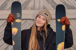A woman in a tan beanie, brown gloves, and a dark jacket smiles while holding Icelantic Skis Tempest 88 skis with yellow flower designs, standing in front of a wooden background.